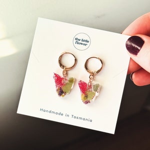 Peut inclure: Boucles d'oreilles créoles dorées avec un pendentif en forme de papillon en résine transparente avec des fleurs séchées et de la verdure incorporées. Les boucles d'oreilles sont faites à la main en Tasmanie. Le texte "the little flower" est imprimé sur la carte.