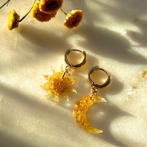 May include: Gold hoop earrings with a sun and moon charm. The charms are made of clear resin with gold flecks.