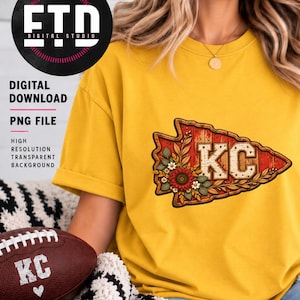 May include: A mustard yellow t-shirt featuring a graphic of a red arrowhead with a wood grain texture, the letters KC, and floral accents. The design includes a sunflower and small white flowers. A brown football with the letters KC is also visible.