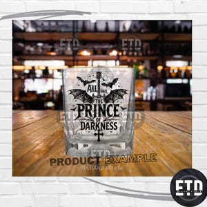 Puede incluir: Un vaso de whisky transparente y cuadrado con el texto "ALL HAIL THE PRINCE OF DARKNESS" en fuente g&oacute;tica, junto con ilustraciones de murci&eacute;lagos y guitarra. El vaso est&aacute; sobre una superficie de madera.
