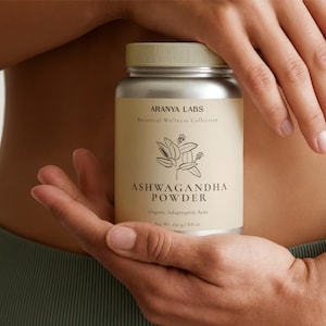 Può includere: Un barattolo d'argento di polvere di Ashwagandha è tenuto in mani aperte. L'etichetta recita "Aranya Labs Botanical Wellness Collection" con un'illustrazione botanica. Il peso netto è di 250 g / 8,8 oz.
