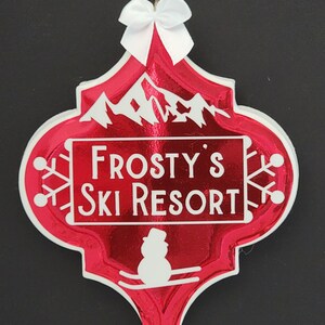 May include: Red and white Christmas ornament shaped like a diamond with a white bow at the top. The ornament has a white silhouette of a snowman skiing down a mountain with the text "Frosty's Ski Resort" on a white banner.