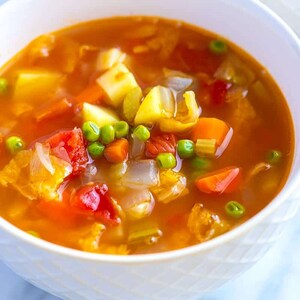 May include: A white bowl filled with a hearty vegetable soup. The soup is a vibrant orange color and contains carrots, potatoes, peas, and other vegetables.
