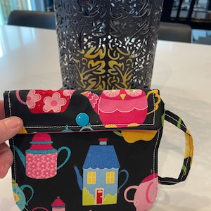 May include: A black fabric pouch with a colorful teacup pattern. The pouch has a flap with a blue button and a handle. The pattern includes teapots, houses, and owls.