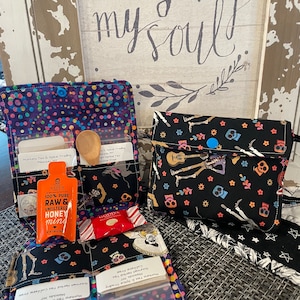 May include: Three small fabric pouches with different patterns. The pouches are filled with small items, including tea bags, a wooden spoon, a honey stick, and a small candy. The pouches are on a woven mat with a sign that says "where my soul sings".