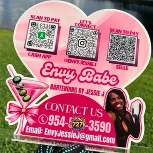 May include: A pink heart-shaped sign with the text "Envy Babe" and "Bartending by Jessie J." It includes QR codes for Cash App, Instagram, and Zelle, along with contact information. A pink martini glass graphic is also present.