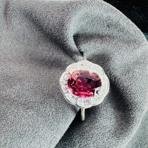 Puede incluir: Un anillo de plata con una gran gema ovalada de color rosa intenso en el centro, rodeada por un halo de piedras más pequeñas y transparentes. El anillo se encuentra sobre un fondo de tela gris, con un vistazo a una caja dorada en la esquina superior derecha.
