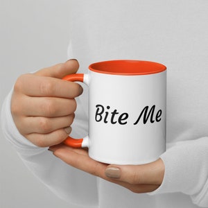 Peut inclure: Une tasse en céramique blanche avec une bordure et une poignée orange. La tasse a le texte "Bite Me" imprimé en cursive noire sur le devant.