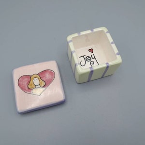 Puede incluir: Una pequeña caja de cerámica con una tapa a rayas de color verde claro y morado. La tapa tiene la palabra "Joy" escrita en negro con un pequeño corazón rojo sobre la palabra. La parte inferior de la caja es rosa con un diseño en forma de corazón y una imagen de dibujos animados de una mujer con cabello rubio dentro del corazón.