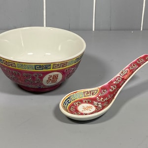 Puede incluir: Un cuenco de porcelana rojo y blanco con un borde dorado y azul y caracteres chinos. El cuenco se acompaña de una cuchara de porcelana roja y blanca a juego.