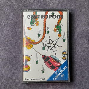 Puede incluir: Un cartucho de juego vintage de Commodore 64 con el juego "Centropods". La carátula presenta un diseño colorido y de dibujos animados con el título del juego, una criatura parecida a un ciempiés y una nave espacial. También se ve el texto "Joystick required".