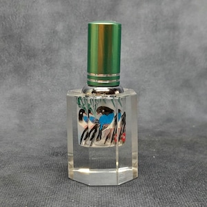 Könnte beinhalten: Ein klarer, facettierter Parfümflakon mit grünem Deckel und silberner Zierleiste. Die Flasche hat ein gemaltes Design von Vögeln mit blauen Flügeln und schwarz-weißen Details. Die Flasche ist rechteckig.