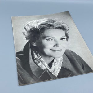 以下が含まれることがあります： 短く波打つ髪をした女性の白黒写真。彼女は暗いジャケットと模様のあるスカーフを着ています。彼女はカメラに向かって微笑んでいます。