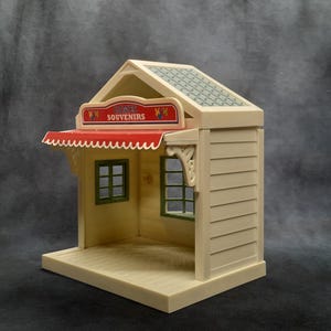 May include: A miniature souvenir shop toy in cream and beige with a red awning. The shop has a sign that reads "Souvenirs" and green window frames. The roof is patterned with a green and gray design. The shop is a toy.