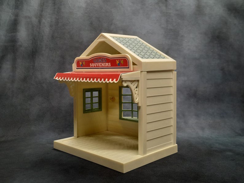 May include: A miniature souvenir shop toy in cream and beige with a red awning. The shop has a sign that reads "Souvenirs" and green window frames. The roof is patterned with a green and gray design. The shop is a toy.