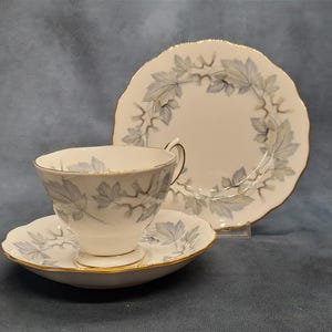 May include: A white teacup, saucer, and plate set with a delicate blue leaf pattern and gold trim. The teacup is on a matching saucer, and a plate is behind them. The set is on a blue background.