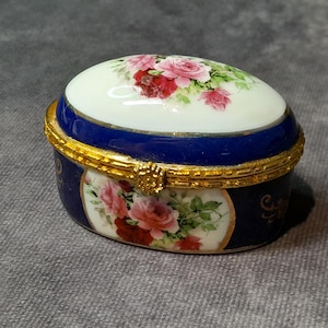 Könnte beinhalten: Ovale Schmuckdose mit weißem Deckel, verziert mit rosa und roten Rosen. Die Box hat ein marineblaues Band und einen Sockel, akzentuiert mit goldenen Zierleisten. Ein Blumendesign befindet sich ebenfalls an den Seiten.