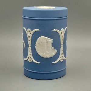 Op de afbeelding: Een blauwe en witte keramische pot met een deksel. De pot heeft een wit ontwerp van een gebouw met een krans eromheen.