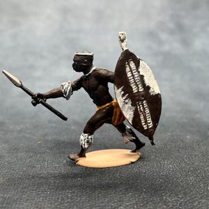 May include: A miniature figurine of an African warrior in a dynamic pose, holding a spear and shield. The warrior is depicted with dark skin, wearing traditional attire, and the shield has white markings. The figurine is on a tan base.
