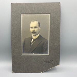 May include: A black and white portrait photograph of a man with a mustache wearing a suit and tie. The photo is framed in a black mat with a white border. The photographer's signature is visible in the bottom right corner.
