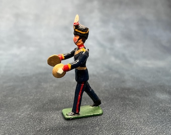 Op de afbeelding: Een miniatuur speelgoedsoldaatfiguur in een marcherende pose, met cimbalen. De figuur is gekleed in een donkerblauw uniform met rode accenten, een zwarte hoed en een groene basis. De cimbalen zijn goudkleurig.