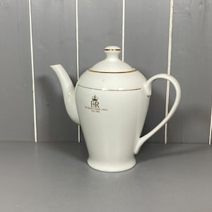 Op de afbeelding: Witte theepot van keramiek met een gouden rand en een gouden embleem met de tekst "ER The Queen's Golden Jubilee 1952-2002".