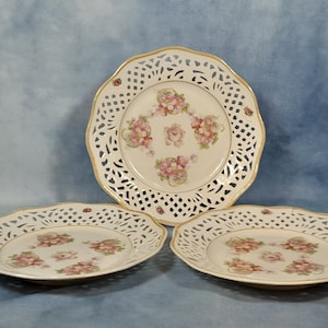 May include: Three white porcelain plates with a delicate openwork design and floral pattern. The plates have a gold trim and feature pink and green roses.