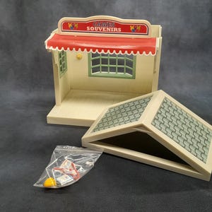 May include: A vintage toy shop playset with a red and white striped awning, green window frames, and a tiled roof. The sign reads "GAMES SOUVENIRS". Includes a clear plastic bag with small accessories.