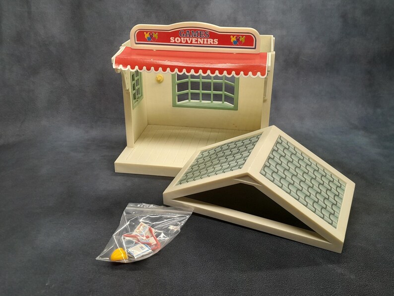 May include: A vintage toy shop playset with a red and white striped awning, green window frames, and a tiled roof. The sign reads "GAMES SOUVENIRS". Includes a clear plastic bag with small accessories.