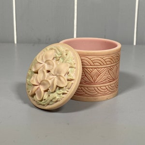 May include: A small, round, pink trinket box with a floral design on the lid. The lid features a carved design of three white flowers with green leaves. The box has a pink and white geometric pattern.
