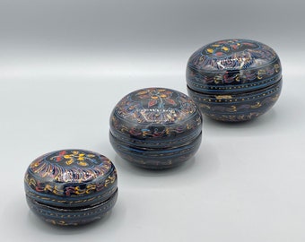 May include: Three round, black, wooden boxes with intricate, colorful floral designs. The boxes are nested inside each other, with the largest box at the top and the smallest box at the bottom.
