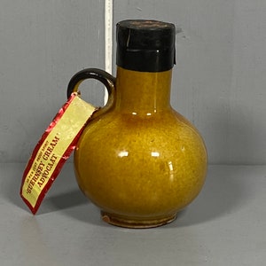 May include: A small brown ceramic jug with a black lid and a red and white label that reads "35% 17.5% Proof Spirit Guernsey Cream Advocaat".