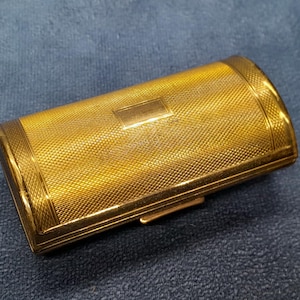 May include: A rectangular, gold-toned metal case with a textured, cross-hatched pattern. The case has a small rectangular accent on the top and a clasp on the side. The edges are smooth and rounded, and the overall design is classic and elegant.