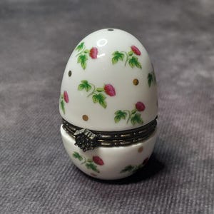 May include: A white, egg-shaped trinket box with a hinged lid. The box is decorated with pink floral patterns, green leaves, and gold dots. A decorative metal clasp secures the lid. The box is likely made of porcelain or ceramic.
