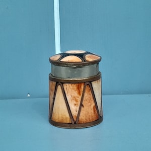 以下が含まれることがあります： 蓋付きの小さな丸い装飾箱。箱は骨と金属でできており、幾何学模様が施されています。蓋は星形のデザインで飾られています。