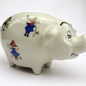 Könnte beinhalten: Weiße Keramik-Sparbüchse mit einem Cartoon-Design von Kindern in blauer und roter Kleidung. Das Sparschwein hat einen Schlitz auf der Rückseite für Münzen.