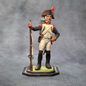 May include: A miniature figurine of a soldier in a historical uniform. The figure wears a black hat with a red plume, a white jacket, and black boots. The soldier holds a brown rifle and stands on a green and black base.