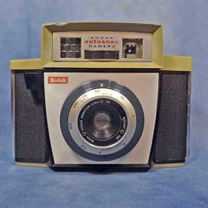Peut inclure: Un appareil photo Kodak Autosnap vintage avec un corps noir et argent. L'appareil photo est recouvert de simili cuir noir à l'avant et à l'arrière. L'objectif est un objectif Kodak Anaston avec une longueur focale de 35 mm. L'appareil photo est doté d'un flash intégré et d'un retardateur.