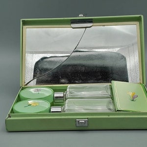 May include: A green metal travel case with a mirror, two glass bottles, two round containers with floral designs, and a green card with a floral design.