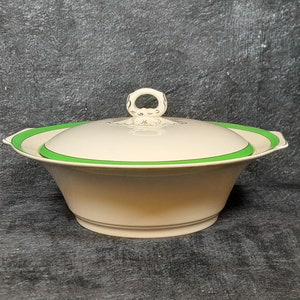 Puede incluir: Un plato de servir de cerámica blanca con un borde verde y una tapa con un asa decorativa. El plato es perfecto para servir una variedad de alimentos.