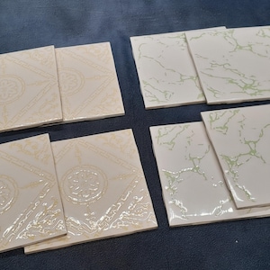 May include: A collection of square ceramic tiles. Some tiles feature a cream-colored floral design with a geometric border. Other tiles have a white marble-like pattern with green veining. The tiles are arranged on a dark blue surface.