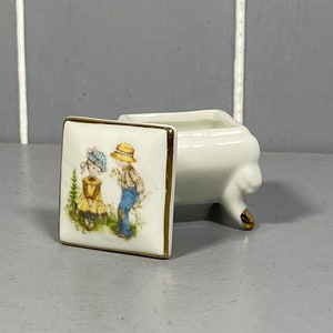 May include: A white porcelain trinket box with gold trim and a square lid featuring a painted image of two children. The box has three gold-painted feet.