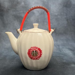 May include: White ceramic teapot with a red and black handle. The teapot has a rounded shape with a lid and a spout. A red and gold emblem is on the front of the teapot. The handle is made of red and black woven material.