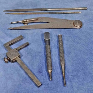 May include: A set of five metal tools, including a pair of dividers, two awls, and two files. The tools are laid out on a blue fabric surface.