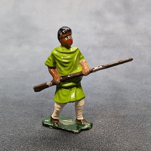 May include: A painted metal toy soldier figure. The figure is dressed in a green tunic, white pants, and brown shoes. It is holding a brown rifle. The figure has black hair and is standing on a green base.