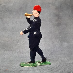 May include: A vintage toy soldier figurine in a dark blue uniform and a red hat, blowing a golden horn. The figure stands on a green base, with details suggesting a historical or military theme.