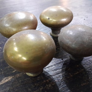 以下が含まれることがあります： 丸みを帯びた楕円形の4つのアンティーク真鍮製キャビネットノブ。ノブは、ヴィンテージであることを示唆する、経年変化と摩耗の兆候を伴う、暖かく金色の色合いをしています。濃い木製の表面に置かれています。