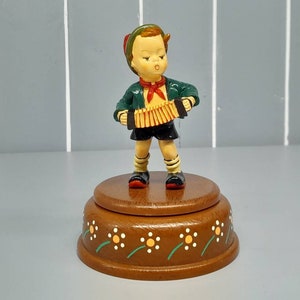 May include: A wooden music box with a figurine of a boy playing an accordion. The boy is wearing a green shirt, red scarf, and black shorts. The music box is decorated with yellow flowers and green leaves.