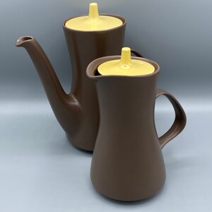 May include: Two-piece brown ceramic coffee set with yellow lids. The set includes a tall coffee pot with a long spout and a smaller pitcher with a handle. The pieces have a matte finish and a minimalist design.