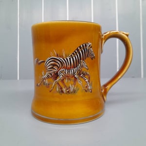 Puede incluir: Una taza de cerámica amarilla con un diseño de cebra en blanco y negro. La taza tiene un asa y una base ancha.